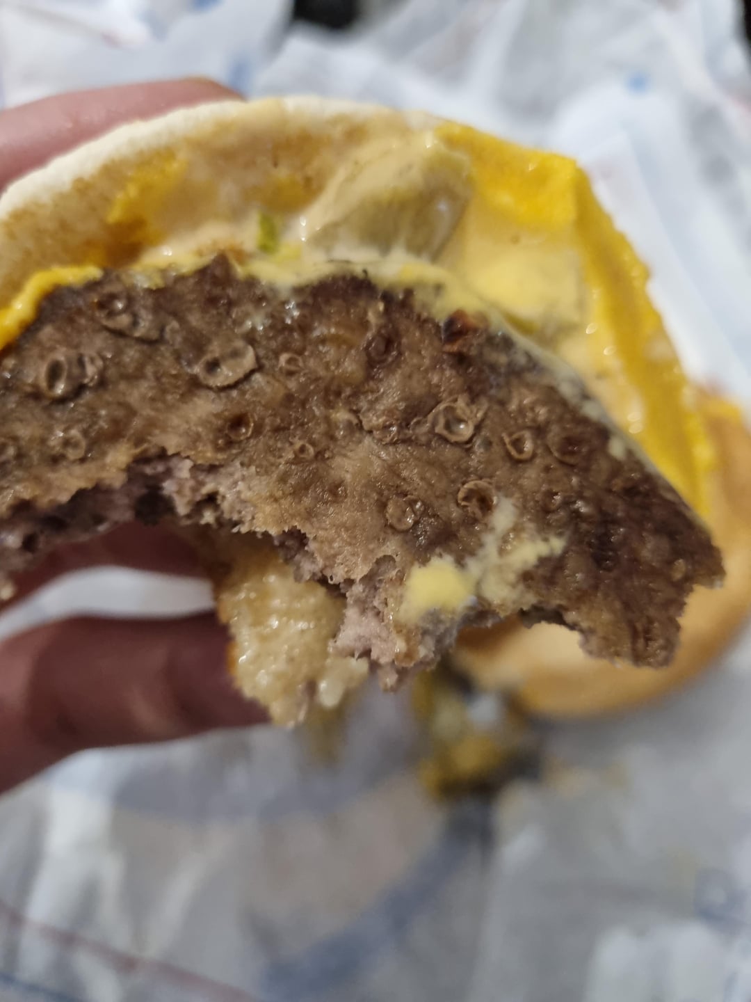 A close-up of a bitten hamburger patty with melted cheese and mustard in a bun, held in someone's hand. The background is slightly blurred, showing a wrapper underneath.