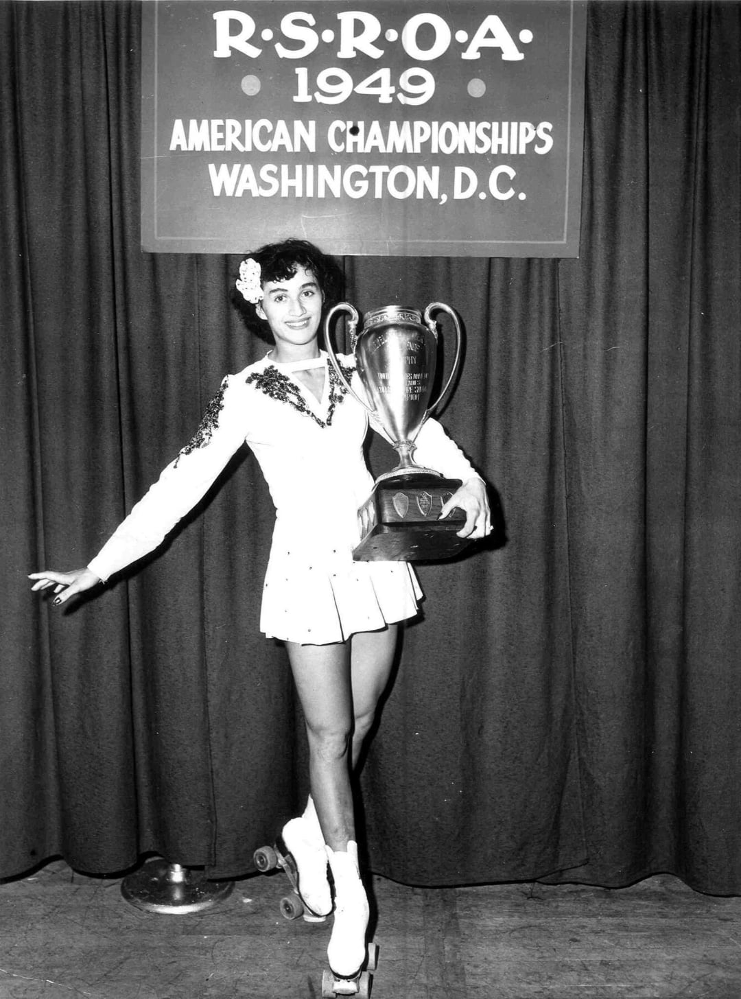 A female roller skater in a costume poses on roller skates, holding a large trophy, in front of a banner reading "R.S.R.O.A. 1949 American Championships Washington, D.C.
