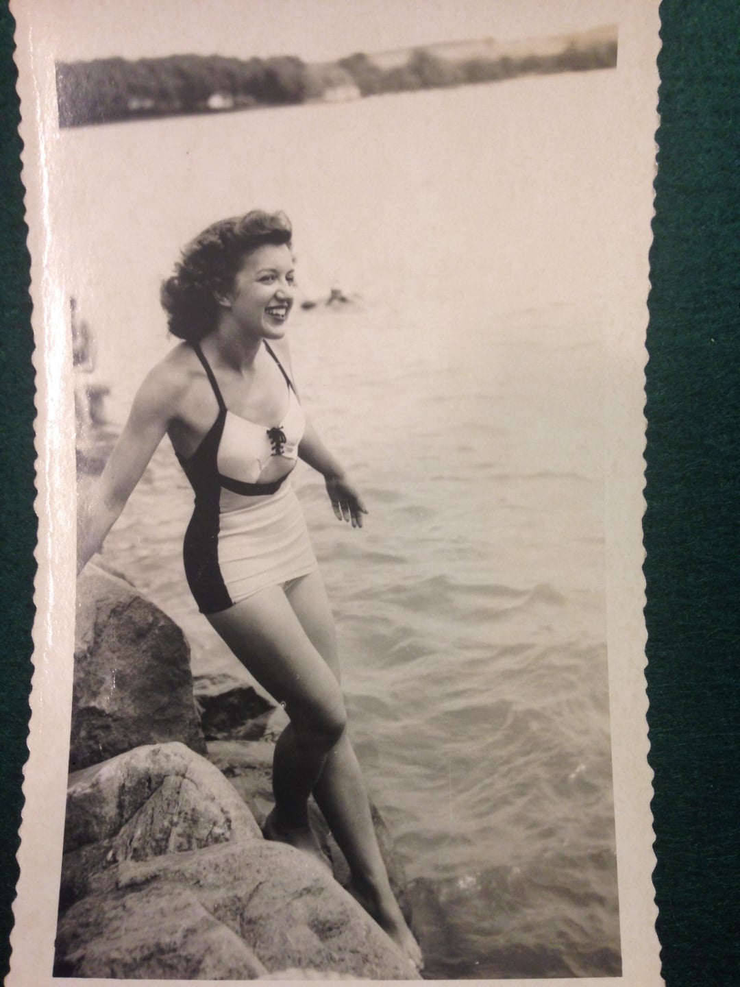 A woman in a vintage swimsuit stands on rocks at the edge of a body of water, smiling and looking to the side. The photo is black and white, and trees are visible across the water in the background.
