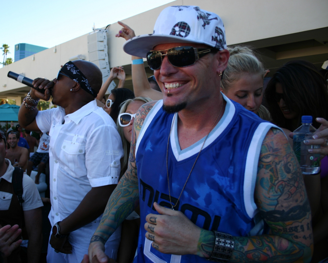 A man in a blue basketball jersey and white cap smiles in a crowded outdoor party. Another man in white sings into a microphone nearby. People around them appear to be enjoying the lively atmosphere.