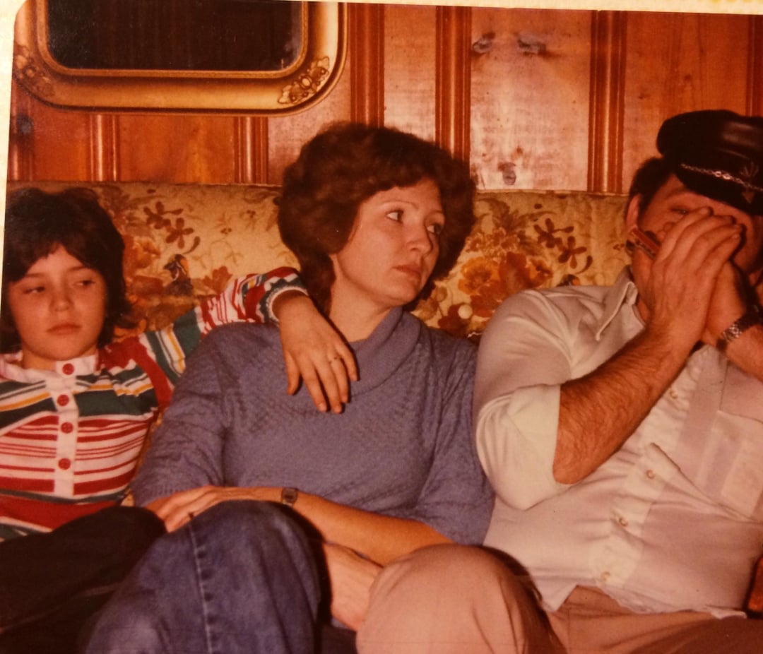 A child, a woman, and a man sit on a patterned couch. The child leans on the woman, who looks at the man. The man, wearing a cap, covers his face with his hands. The setting features wood-paneled walls.