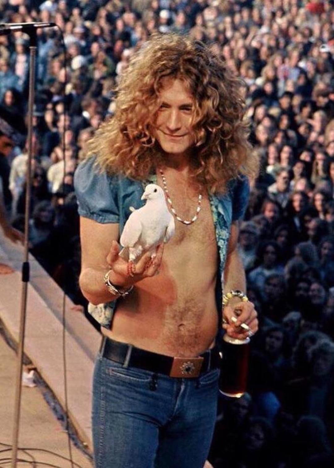 A man with long curly hair, wearing an open shirt and jeans, stands on stage holding a white dove in one hand and a drink in the other, with a microphone and a large crowd in the background.