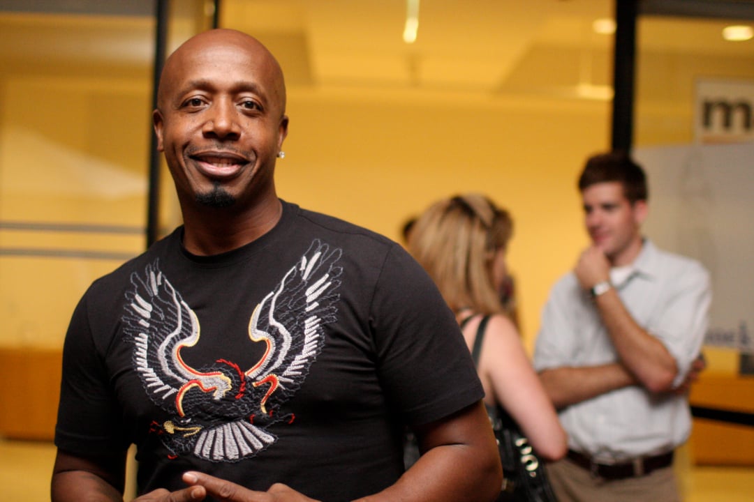 A man in a black t-shirt with a bird graphic stands smiling indoors, with three people talking in the blurred background.