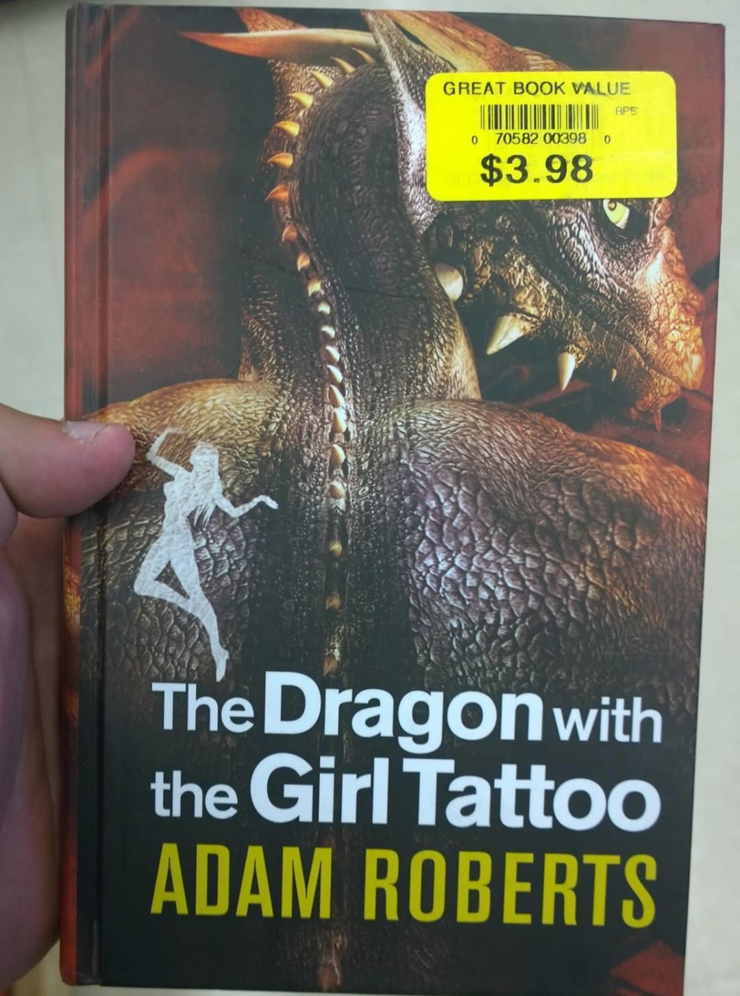 A hand holds a book titled "The Dragon with the Girl Tattoo" by Adam Roberts. The cover shows a dragon’s face and a silhouette of a woman, with a yellow price sticker reading "$3.98 Great Book Value.