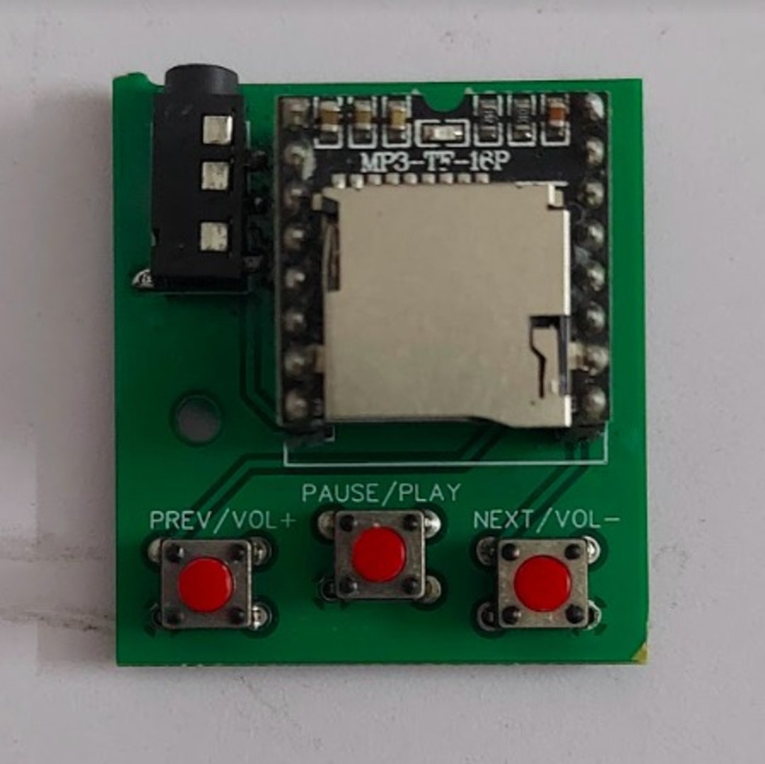A small green circuit board with a microSD card slot, a headphone jack, and three red buttons labeled "PREV/VOL+", "PAUSE/PLAY", and "NEXT/VOL-".
