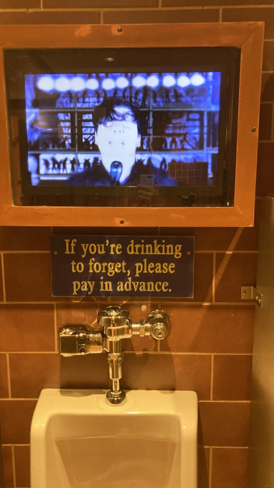 A urinal is mounted below a humorous sign that reads, "If you're drinking to forget, please pay in advance," with a TV screen playing a show above it. The wall is tiled with brown tiles.