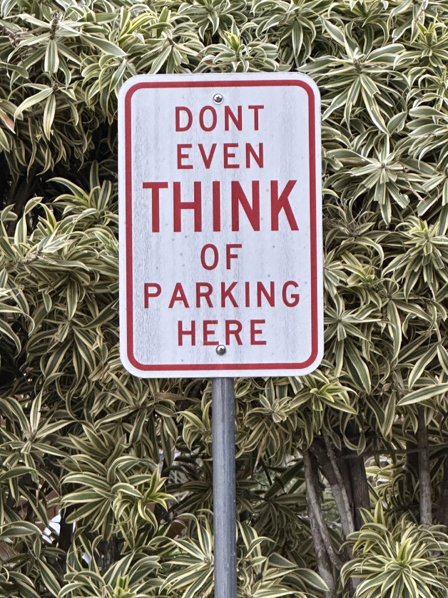 A sign with red text on a white background reads, "DON'T EVEN THINK OF PARKING HERE," in front of leafy green plants.