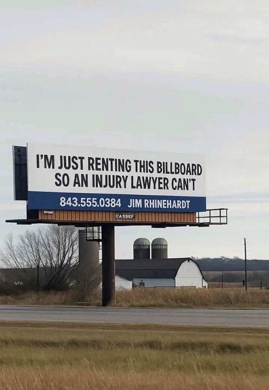 A billboard beside a rural road reads, "I'M JUST RENTING THIS BILLBOARD SO AN INJURY LAWYER CAN'T." Below is a phone number and the name "JIM RHINEHARDT." Farm buildings are visible in the background.