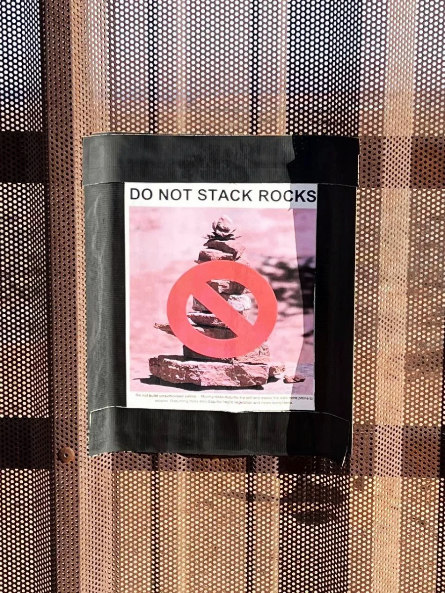 A sign taped to a perforated metal surface shows a photo of stacked rocks with a red prohibition symbol over it and text that reads "DO NOT STACK ROCKS.