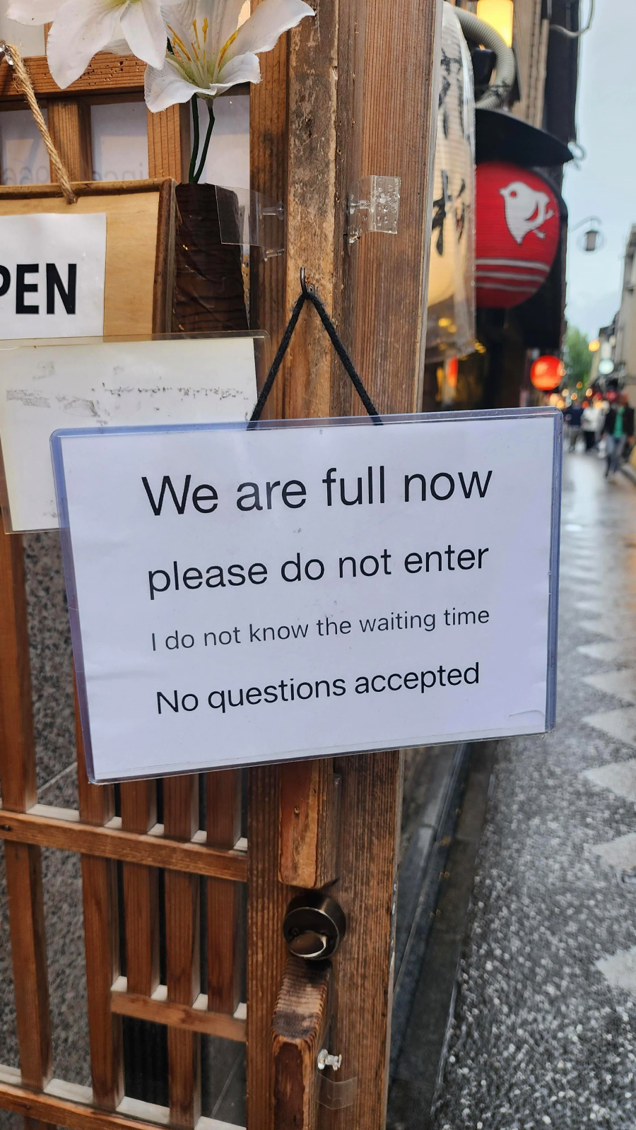 A sign on a wooden door reads: "We are full now please do not enter. I do not know the waiting time. No questions accepted." The background shows a rainy street scene.
