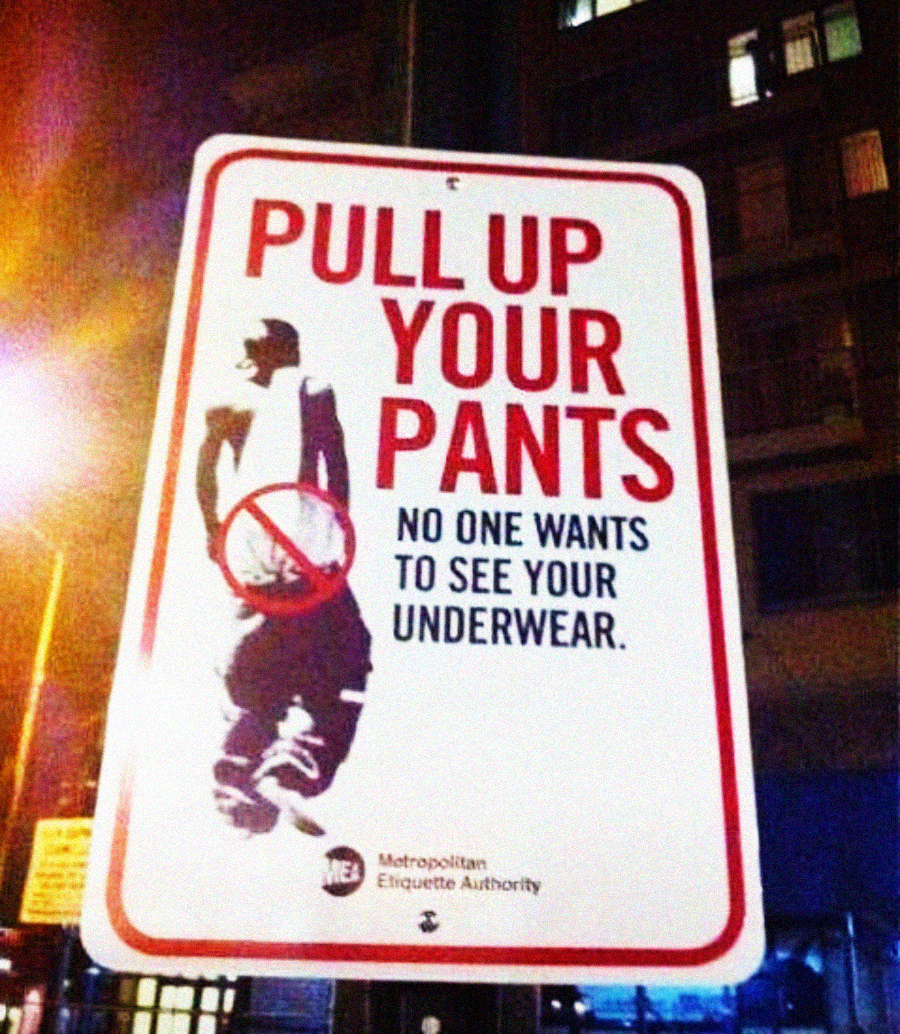 A street sign reads, "PULL UP YOUR PANTS. NO ONE WANTS TO SEE YOUR UNDERWEAR," with an image of a person sagging their pants, crossed out in red, against a city background at night.