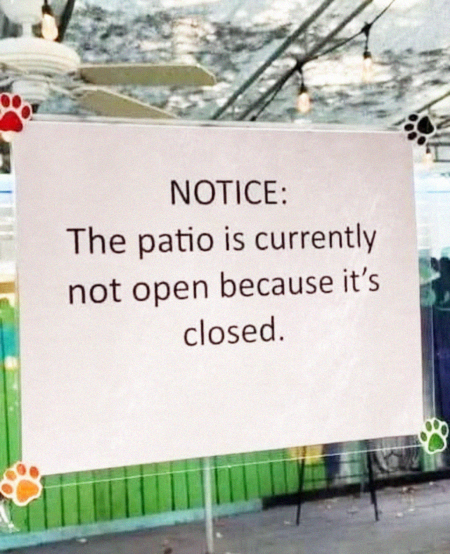A sign reads: “NOTICE: The patio is currently not open because it’s closed.” The sign is decorated with colorful paw prints on the corners and is displayed in front of a fenced outdoor area.