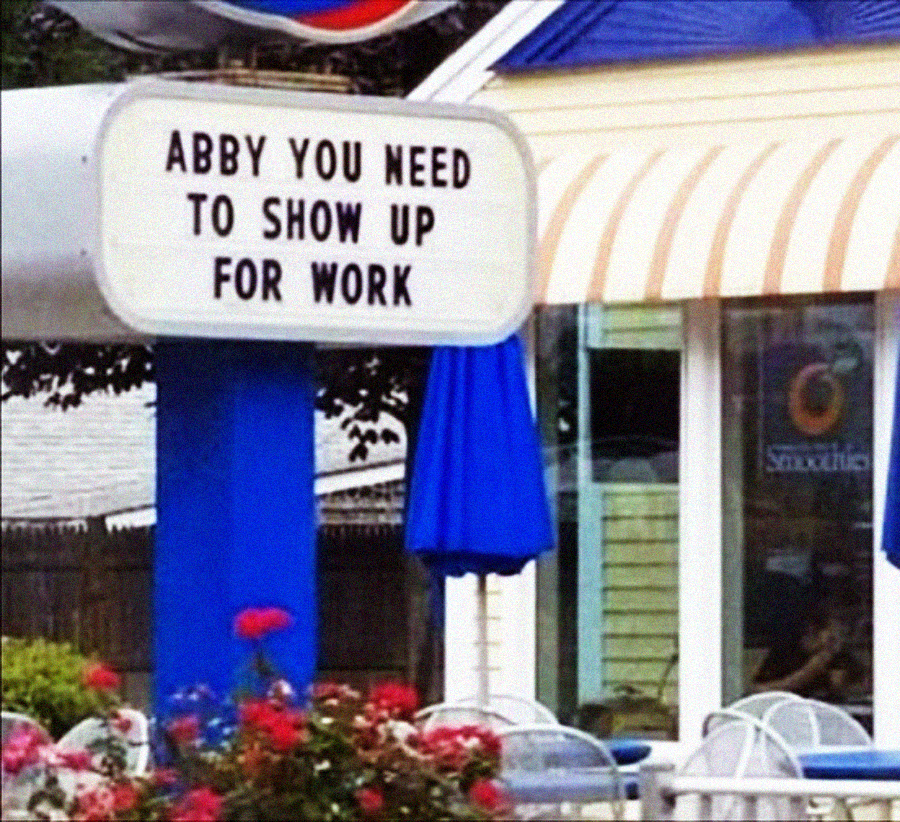 A sign outside a restaurant displays the message, "Abby you need to show up for work." The background shows part of the restaurant building with a blue and yellow awning.