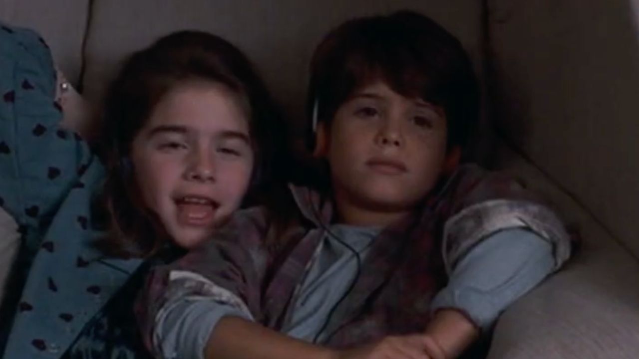 Two children sit close together on a couch. The girl on the left is smiling with her mouth slightly open, while the boy on the right is looking forward with a neutral expression. Both appear relaxed and comfortable.