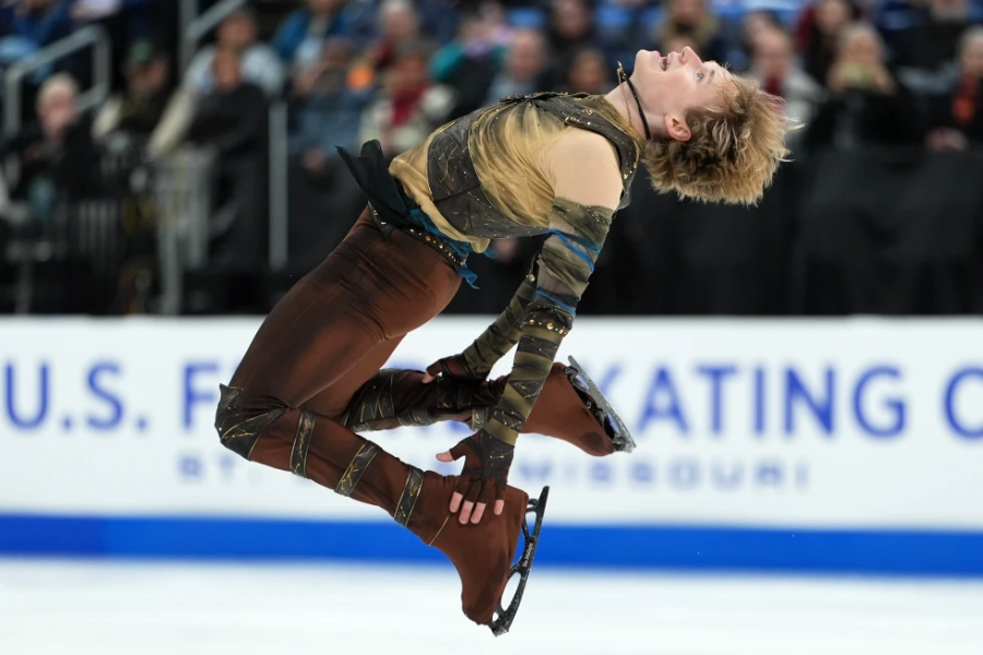A figure skater in a brown and gold costume performs a dramatic jump on the ice, arching his back with knees bent and arms extended, during a competition. Spectators are visible in the background.