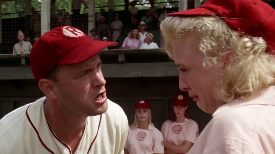 A baseball coach in a red cap angrily yells at a blonde female player in uniform, who appears to be crying. Other female players and spectators watch in the background.