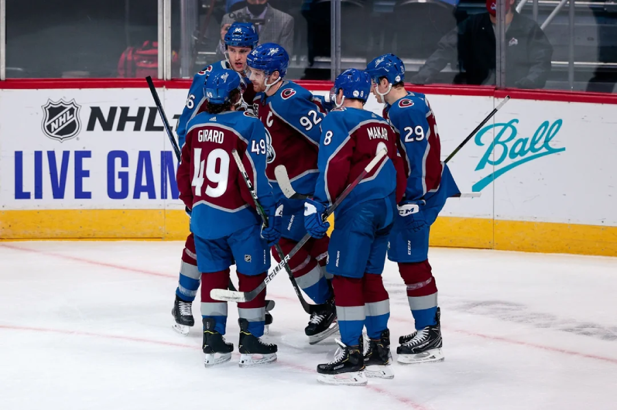 Five Colorado Avalanche hockey players in maroon and blue uniforms huddle together on the ice, appearing to celebrate during an NHL game. The rink boards display NHL and sponsor logos in the background.