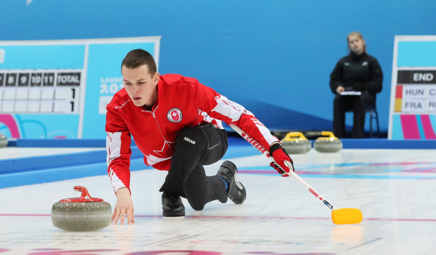 A male curler in a red jacket releases a curling stone on the ice, holding a broom. A scoreboard and another person sitting in the background are visible.