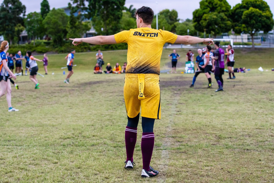 A person in a yellow sports uniform labeled “SNITCH” stands on a grassy field with arms outstretched, surrounded by players in various colored uniforms during an outdoor game.