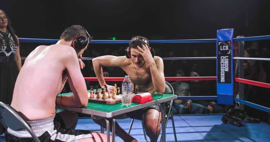 Two men wearing boxing shorts and headphones play chess at a table inside a boxing ring, with an audience watching and a sign reading "chessboxing" on the ropes. Bottled water and a chess clock are on the table.
