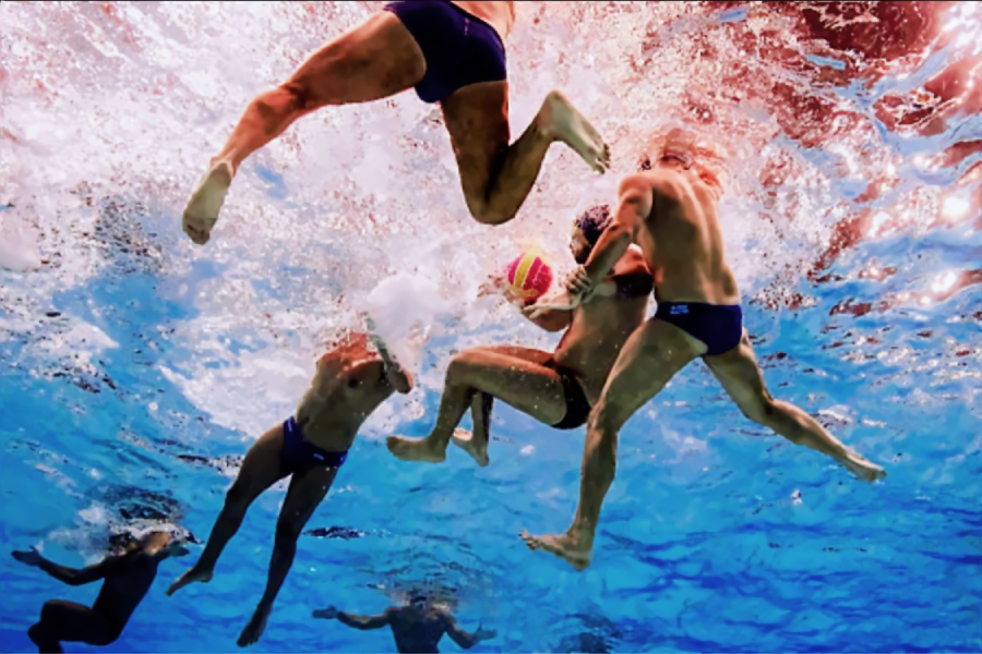 Underwater view of water polo players in swimwear competing for the ball, with splashes and movement visible above and below the water’s surface.