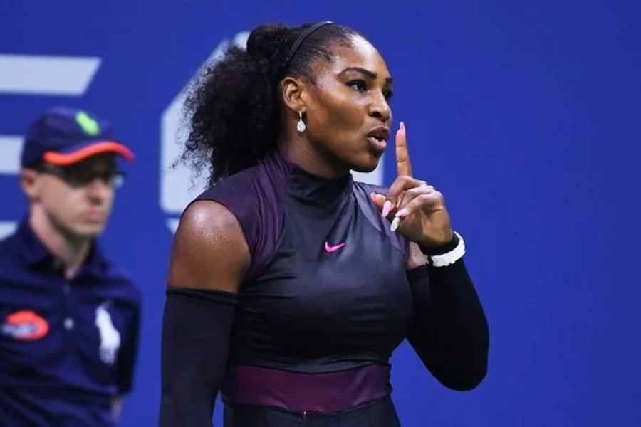 A tennis player in a black and purple outfit gestures with her index finger to her lips, signaling for silence. An official wearing a cap and sunglasses stands in the background.