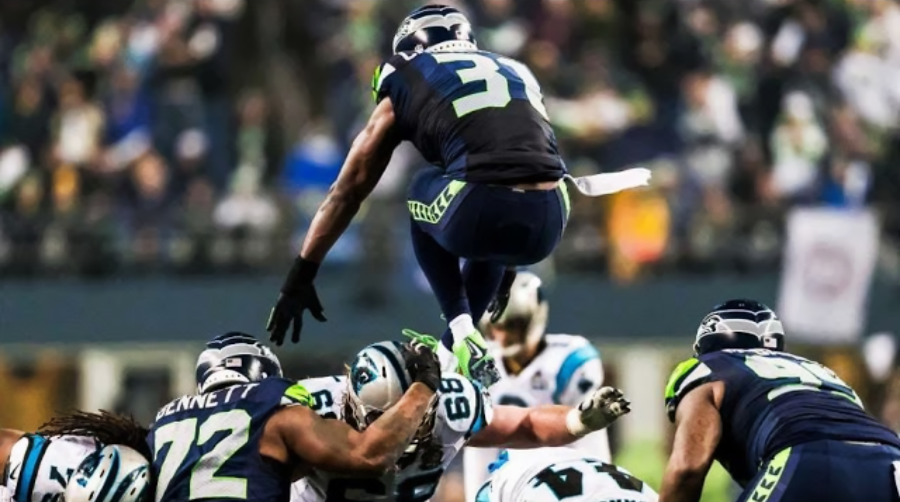 A Seattle Seahawks football player in a dark uniform leaps high over a group of players during an NFL game, with opponents from the Carolina Panthers attempting to block.