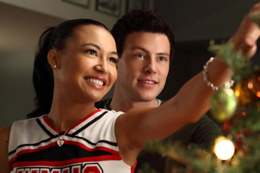 A smiling woman in a cheerleading uniform decorates a Christmas tree, while a man stands behind her, looking on with a gentle smile. The scene is warm and festive.