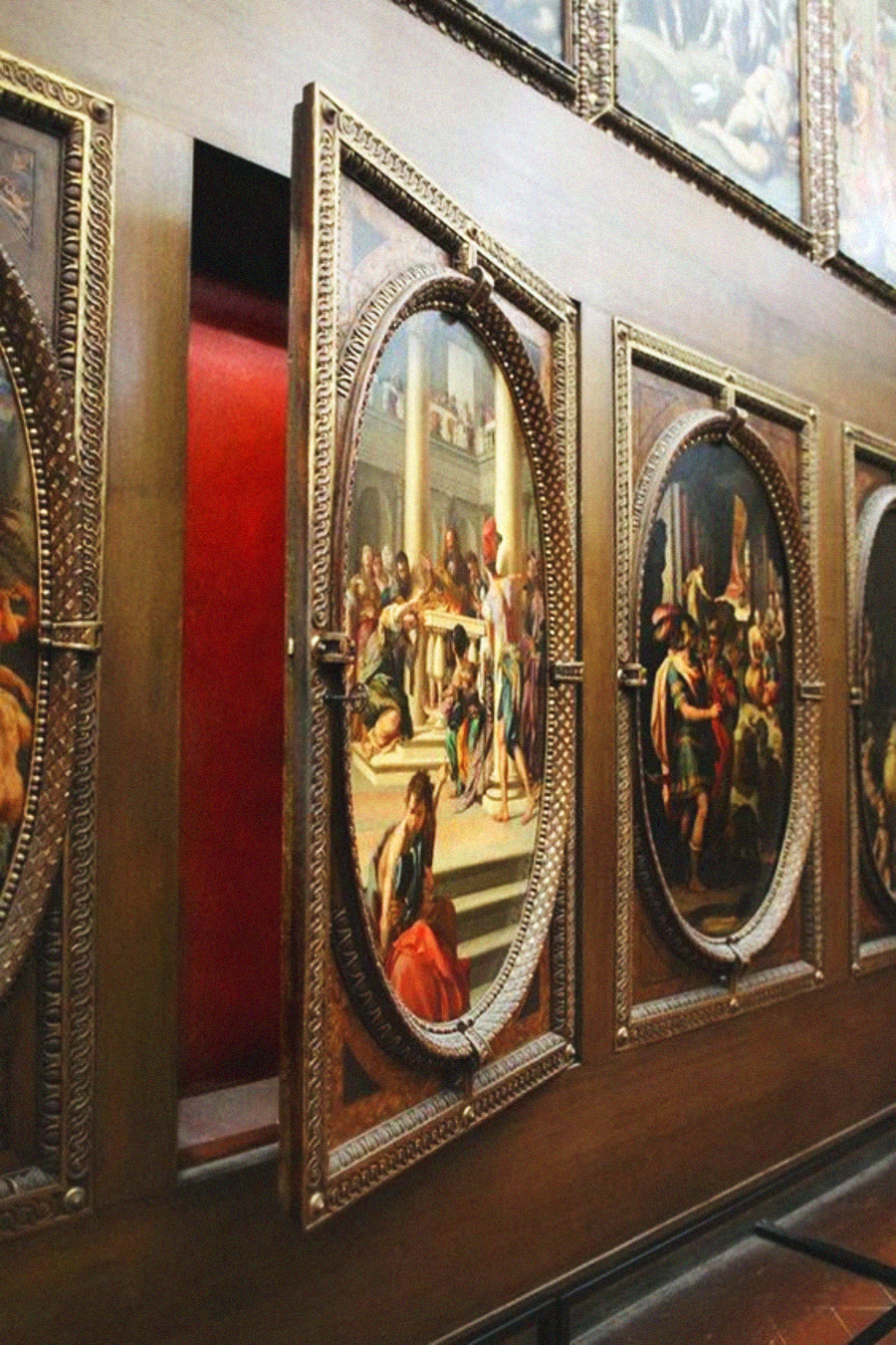 A wooden wall with framed, oval Renaissance paintings, one of which is open like a door to reveal a red interior, suggesting a hidden compartment or secret passage behind the artwork.