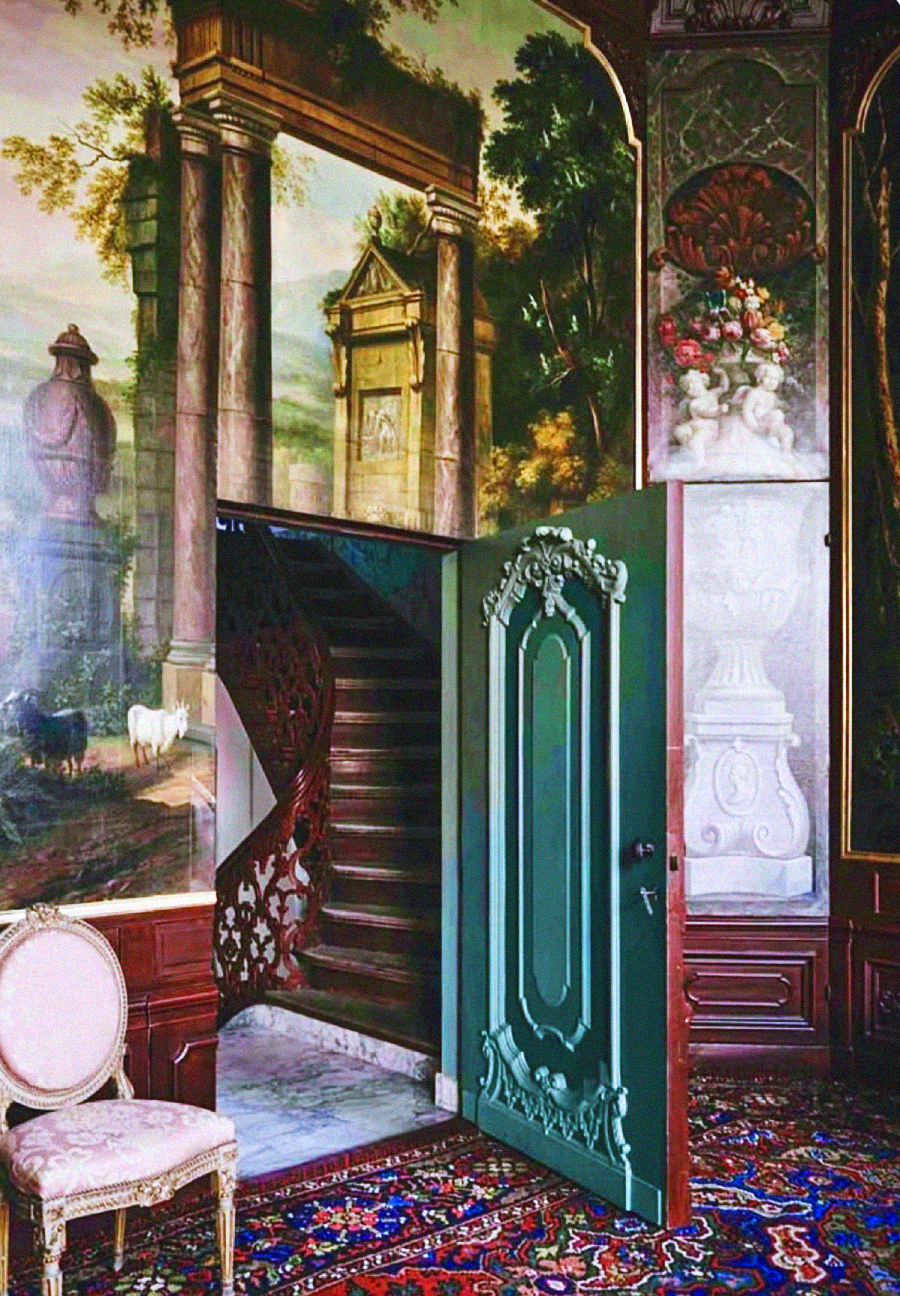 An ornate room with a painted mural of classical ruins and landscapes, an open green door revealing a spiral wooden staircase, an upholstered chair, and an intricate patterned carpet.