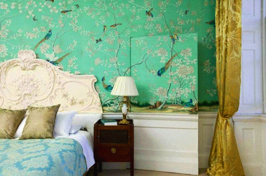 A bedroom with a white ornate headboard, teal bedding, floral and bird-patterned green wallpaper, a wooden nightstand with a lamp, and gold curtains beside a large window.