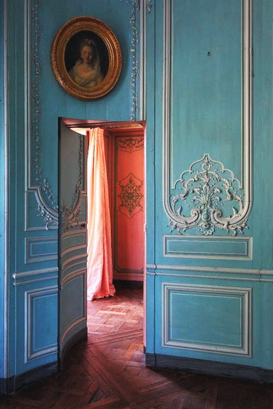 Elegant blue paneled wall with ornate white trim, a small door partly open to a pink room, and a framed oval portrait of a woman above the door. The floors are dark wood in a herringbone pattern.