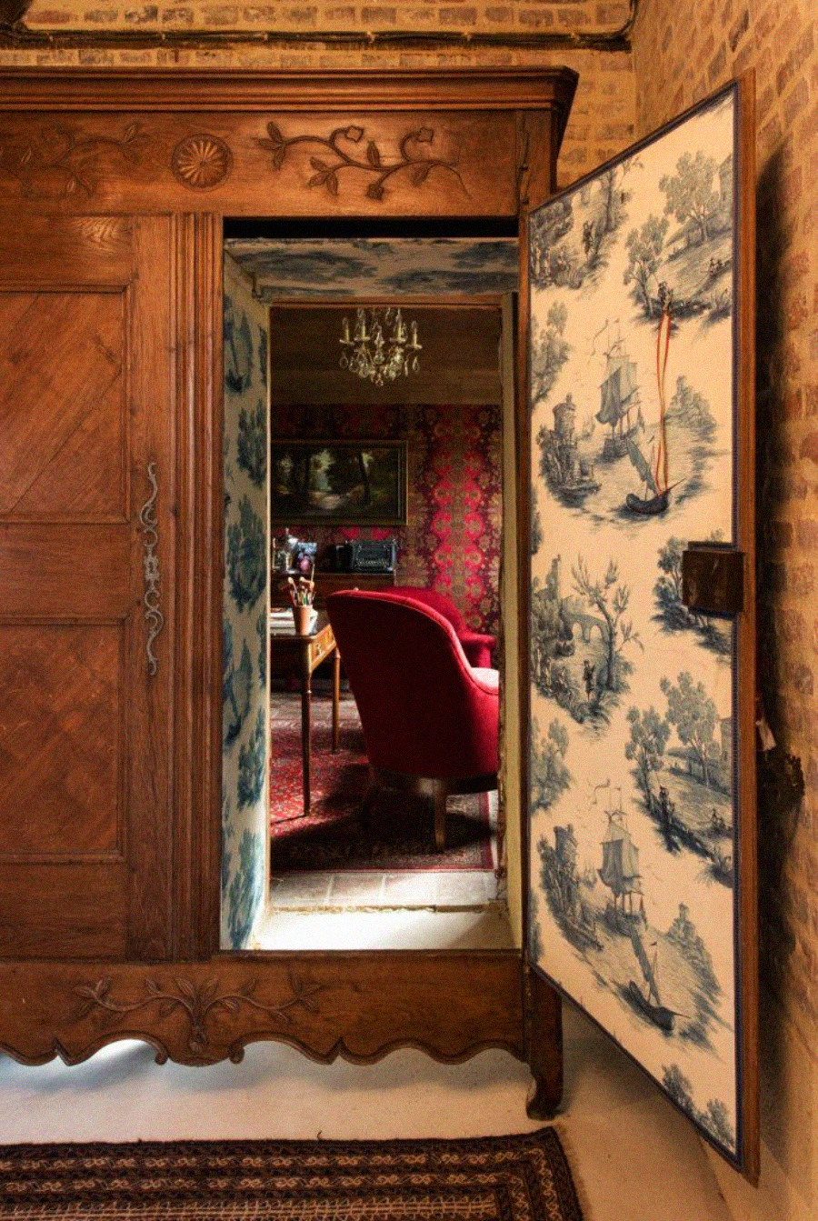 An ornate wooden wardrobe with one door open, revealing a hidden room with vintage decor, red chairs, patterned walls, and a chandelier, contrasting with the wardrobe’s blue-and-white toile lining.