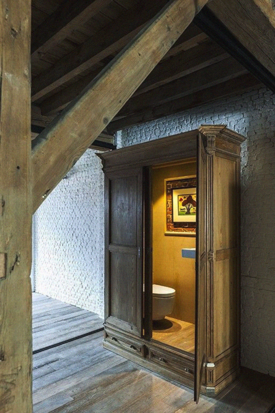 A wooden wardrobe under a staircase opens to reveal a hidden toilet inside, with a framed painting on the back wall and ambient lighting illuminating the small space.