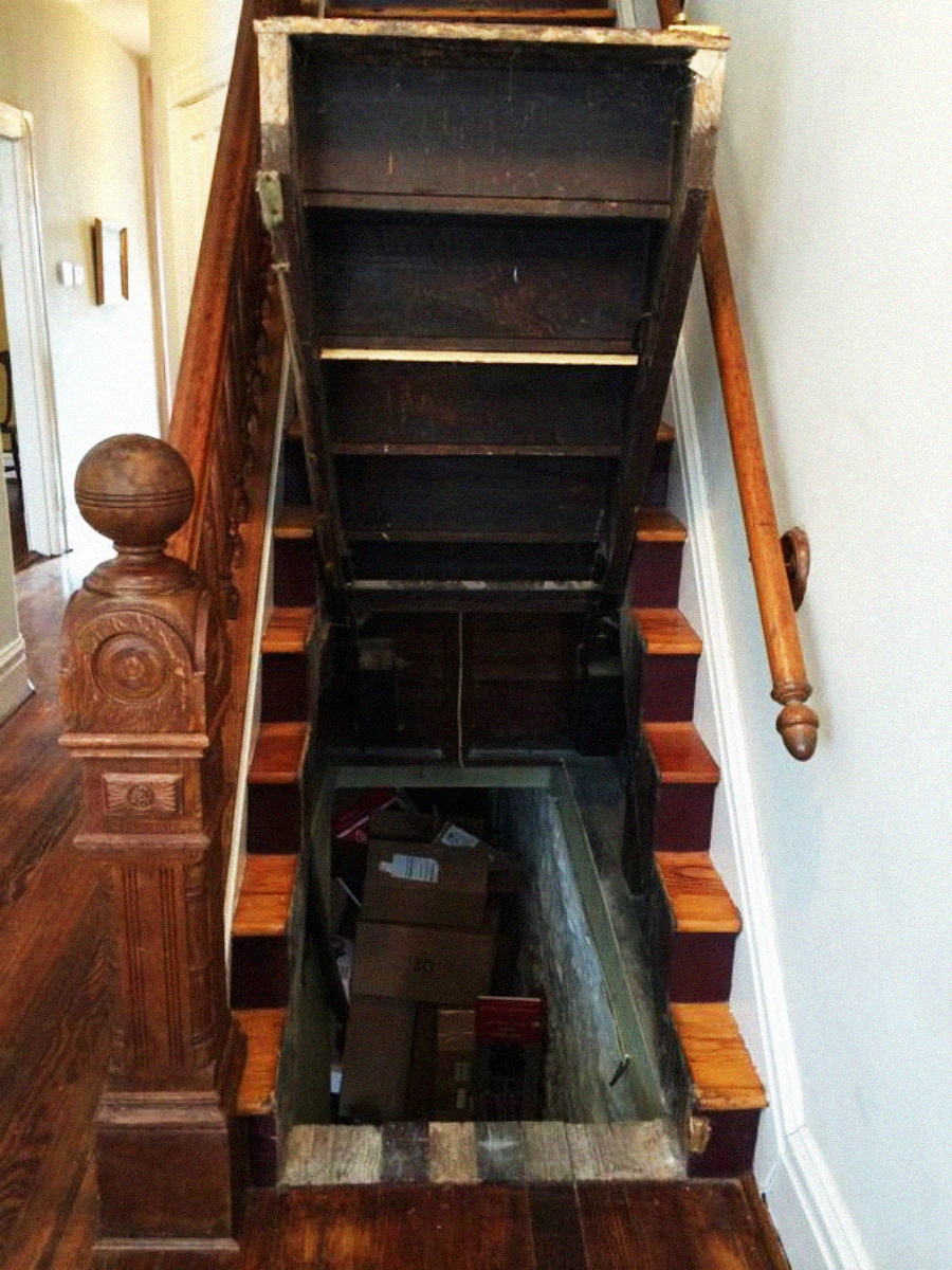 Wooden staircase with the bottom steps lifted up to reveal a hidden storage space underneath, containing several cardboard boxes. The banister and newel post are ornately carved, and the hallway is visible in the background.