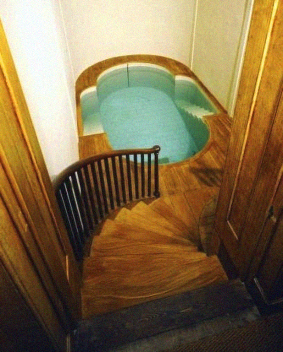 A small, round indoor pool with clear water is surrounded by wooden flooring. Curved wooden steps with a railing lead down to the pool from a doorway.