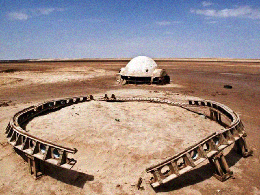 A dome-shaped, weathered structure and a circular metal frame sit abandoned on a flat, barren desert landscape under a blue sky with scattered clouds.