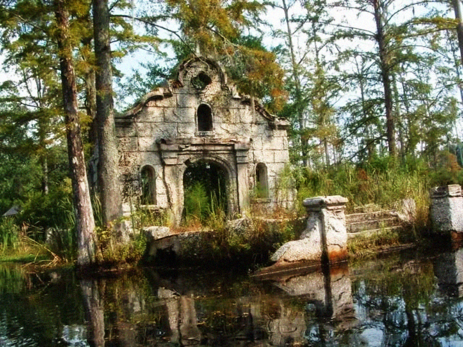 An old, weathered stone ruin with arched windows stands surrounded by tall trees and overgrown plants beside a calm, reflective pond. The scene looks abandoned and serene.