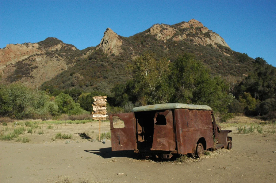 A rusty, abandoned van sits on dry ground with open doors; behind it is a wooden signpost with multiple direction signs, set against a backdrop of green trees and rugged mountains under a clear blue sky.