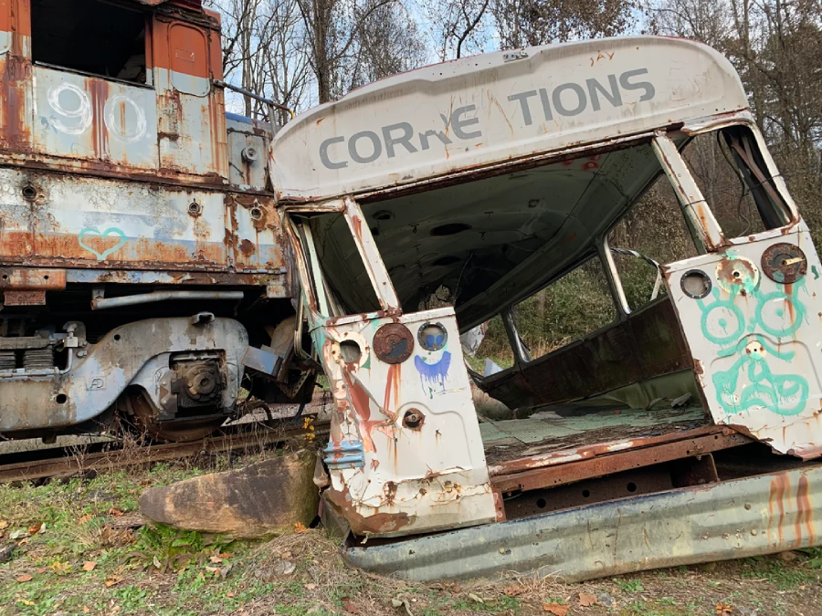 A rusty, damaged bus marked “CORRECTIONS” lies tilted on the ground next to an old, rusted train car. Both vehicles are covered in graffiti and surrounded by grass and trees.