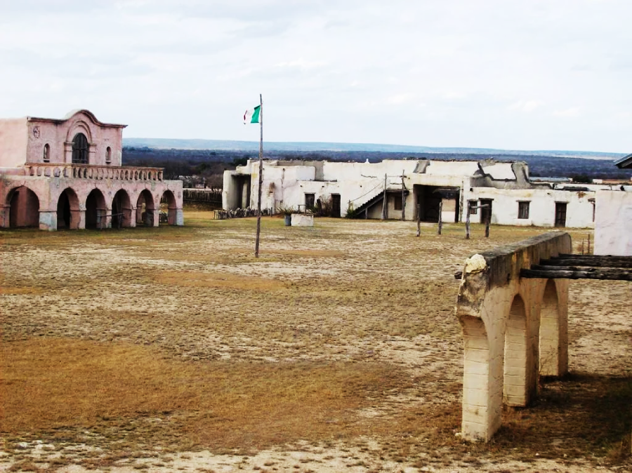 A wide view of an old, weathered fort with arched structures, a central flagpole flying the Mexican flag, and open grassy grounds under a cloudy sky.