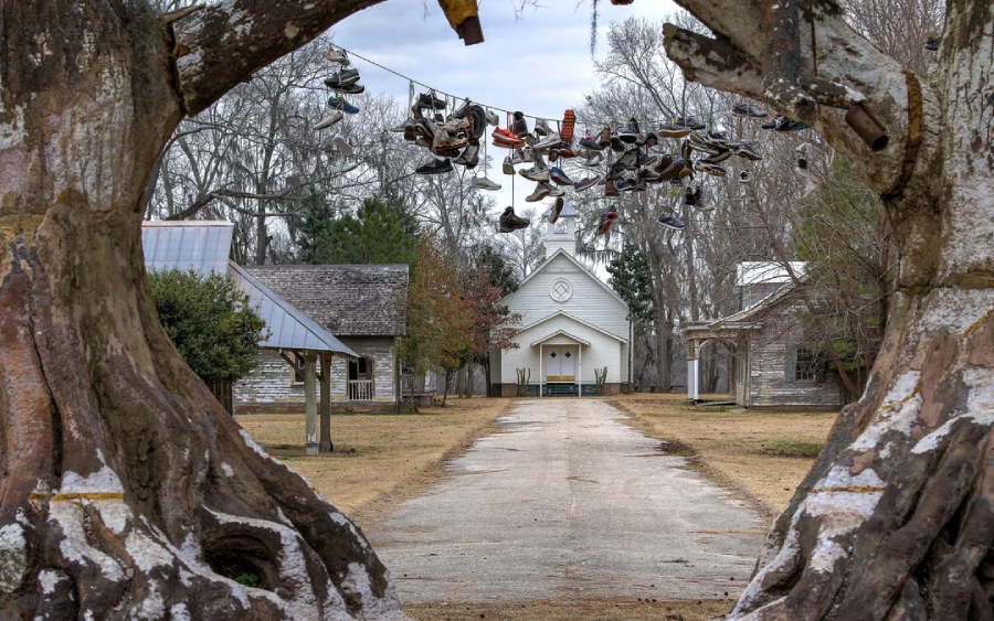 A path lined with old buildings leads to a white church. Shoes hang from a wire suspended between the split trunks of two large trees framing the entrance. The scene appears quiet and overcast.