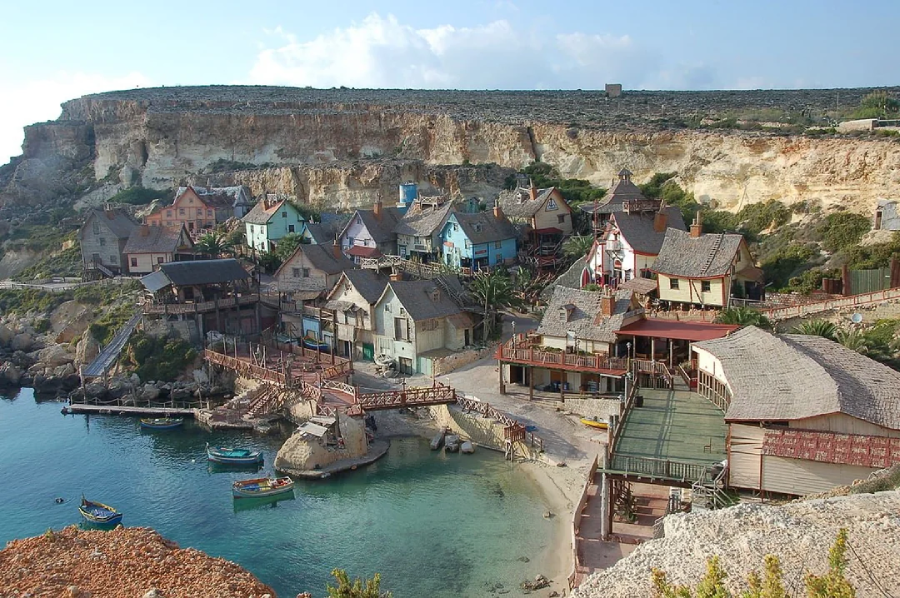 A colorful village with wooden houses sits by a clear blue bay, surrounded by rocky cliffs and greenery. Small boats float in the water, and wooden walkways connect the buildings.