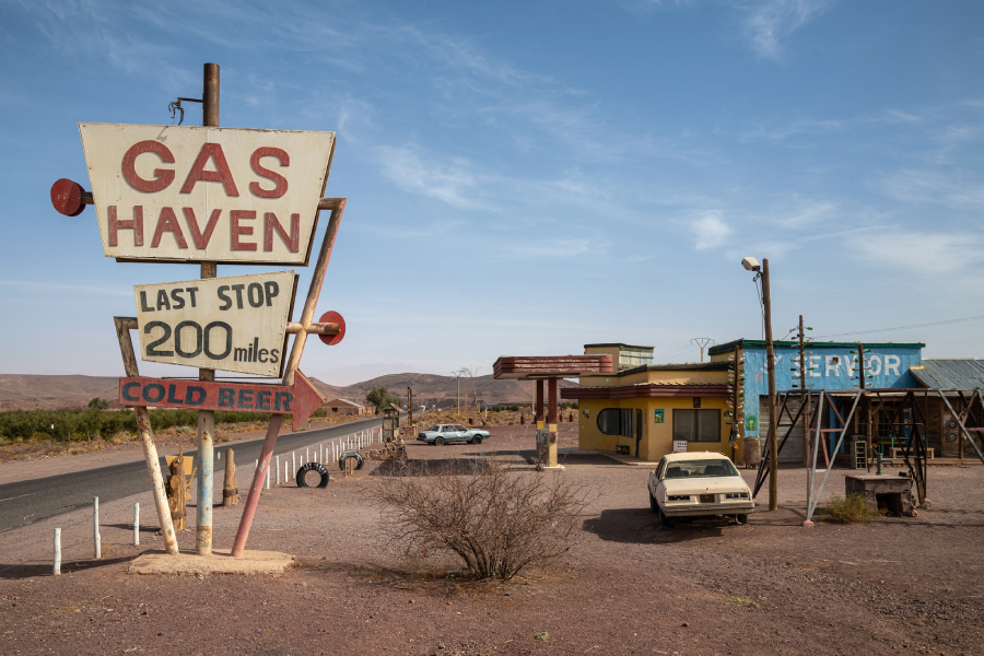 A vintage gas station with a large “Gas Haven” sign reading “Last stop 200 miles, cold beer,” stands beside a deserted road in a barren, desert landscape. Two old cars are parked near the small buildings.
