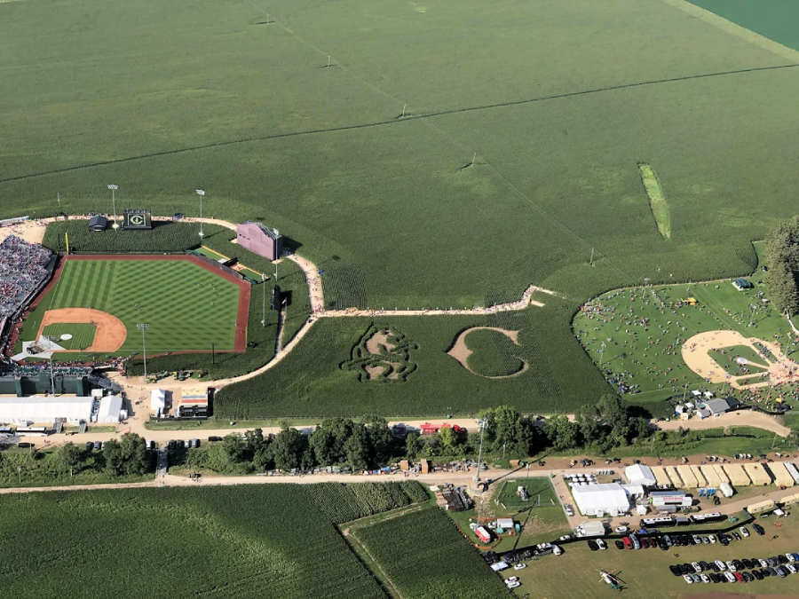 Aerial view of a baseball field surrounded by green cornfields, with a second smaller diamond nearby and a path cutting through the corn. A carved design of a baseball player and logo are visible in the field. Spectators are present.
