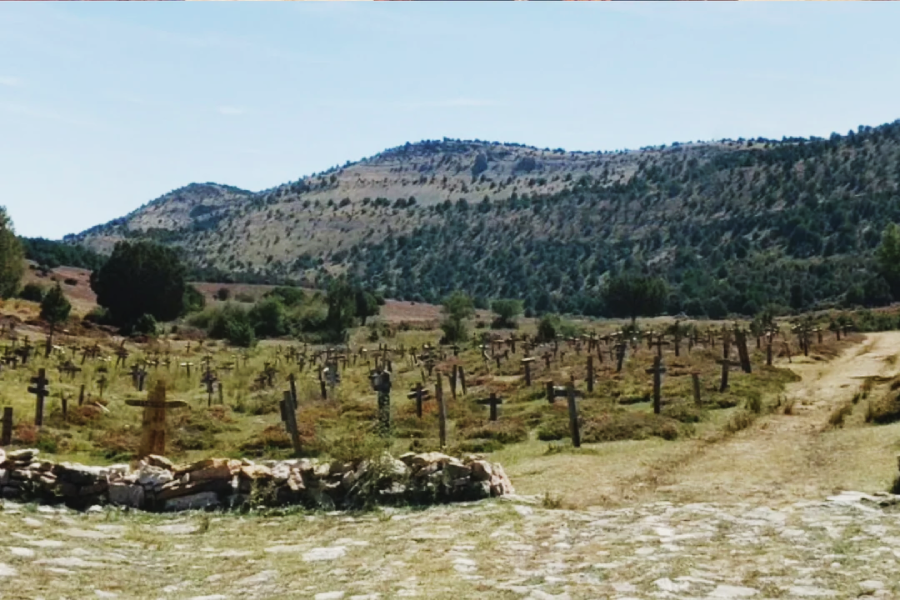 A cemetery with numerous wooden crosses scattered across a grassy field, with hills and trees in the background under a clear sky. A dirt path runs alongside the graves.