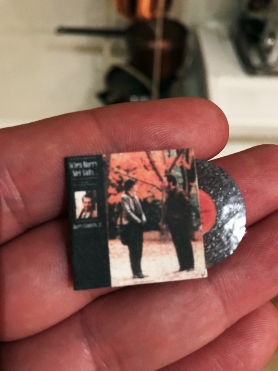A close-up of a hand holding a miniature vinyl record and album cover featuring two men standing and talking outdoors with autumn foliage in the background. The tiny record and cover fit entirely on the person's palm.