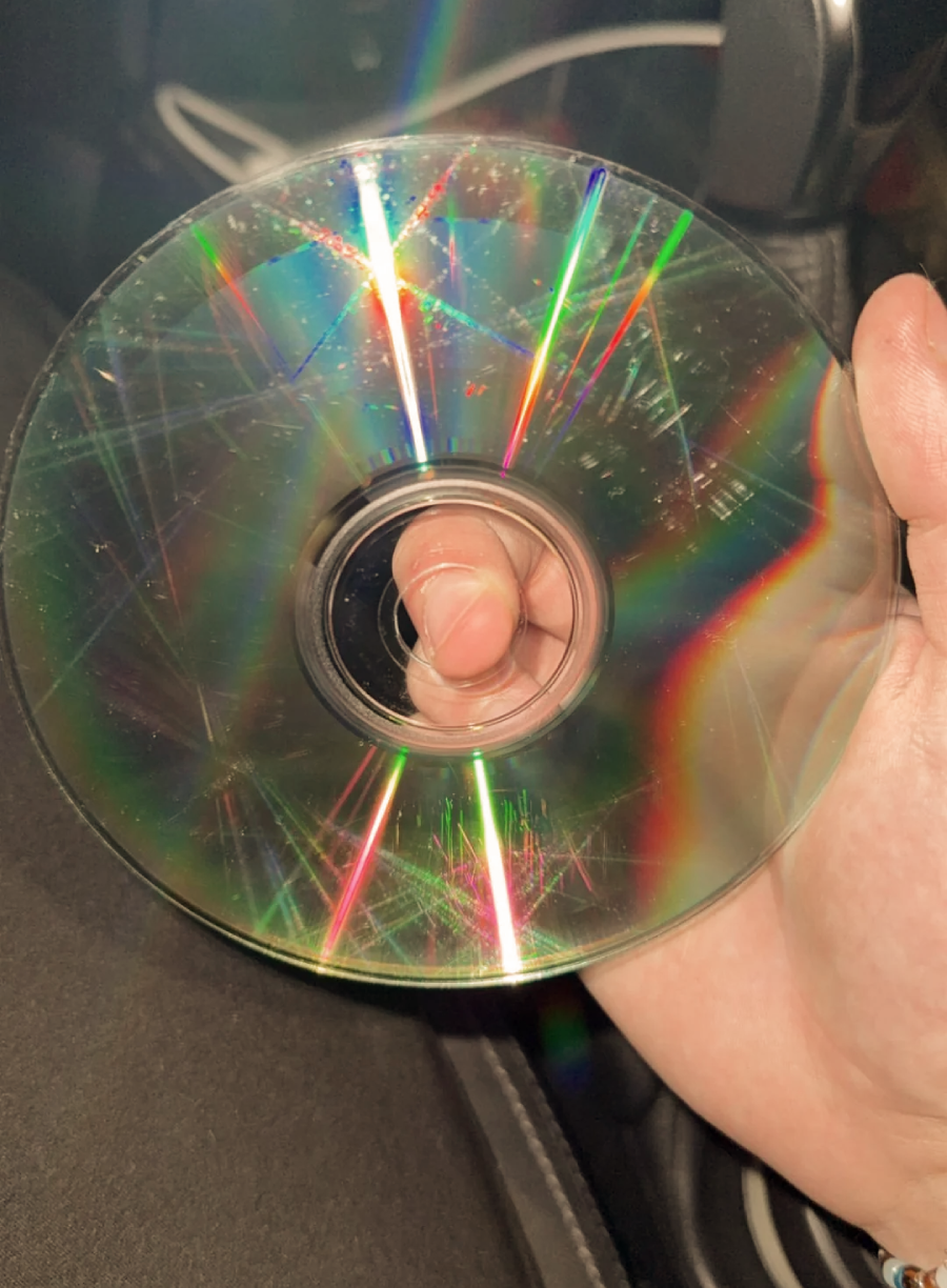 A hand holding a scratched and worn CD with visible colorful light reflections and multiple surface scratches, photographed indoors with a bright flash.