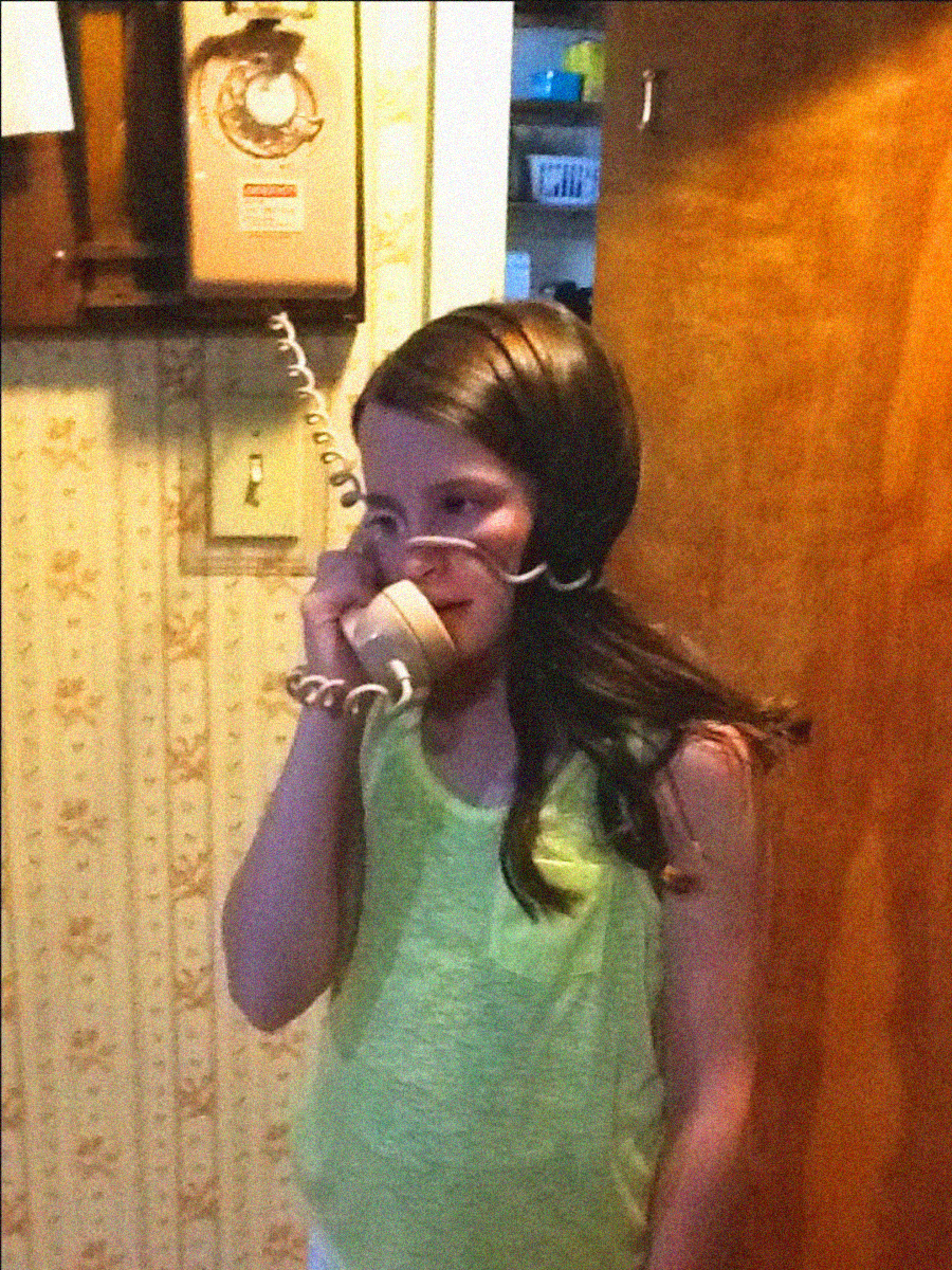 A girl with long dark hair and a yellow tank top holds a beige rotary phone to her ear, standing in a retro-style room with patterned wallpaper and wood paneling. The phone cord is twisted around her head.
