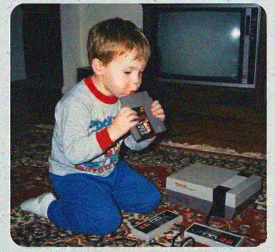 A young boy in pajamas kneels on a patterned rug, holding and blowing into a Nintendo game cartridge in front of an old television and NES console. Two controllers are on the floor beside him.